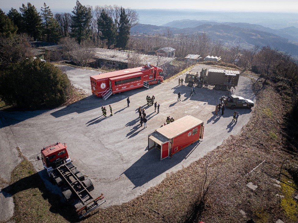 vista dall'alto dei mezzi e laboratori mobili stazionati in un piazzale sopraelevato con vista di sfondo di paesaggio collinare
